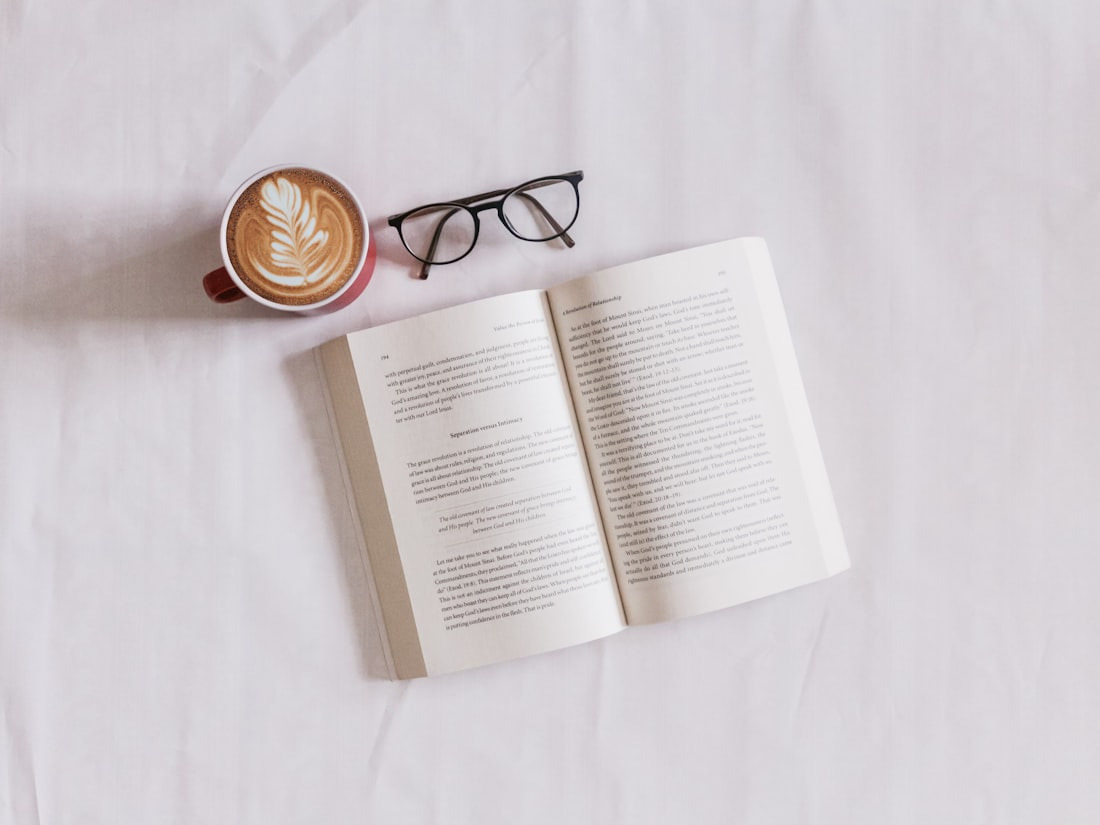 Secangkir kopi hangat di samping kacamata baca dan buku terbuka dalam cahaya pagi yang lembut
